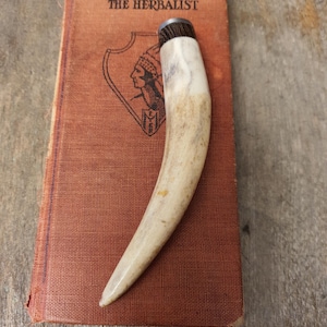 May include: A curved, off-white antler handle with a dark brown, textured wood cap and a silver-toned metal ferrule, resting on a rectangular, orange-brown book titled THE HERBALIST with a black line drawing of a person in a shield emblem.
