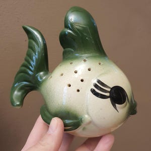 May include: A ceramic fish-shaped shaker featuring a glossy cream-colored body with dark green gradient accents on the fins and tail. The top of the fish has a series of small circular holes, and the face is decorated with painted black eyelashes.