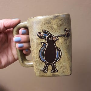 May include: A ceramic mug with a speckled tan glaze featuring a dark brown, stylized bird illustration on the front. The bird has blue wing accents and is depicted with a long beak holding a small leaf. The mug has a sturdy, rectangular handle.