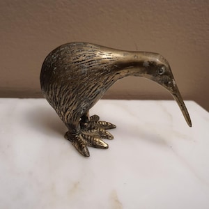 May include: A brass kiwi bird figurine featuring a textured body that mimics feathers, a long downward-curving beak, and detailed feet. The metal surface displays a weathered, aged patina with dark brown and gold tones throughout the sculpture.