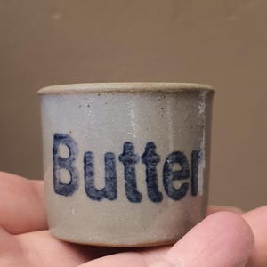May include: A small, cylindrical stoneware butter dish with a light gray exterior and a darker gray rim. The word "Butter" is stenciled in dark blue on the front. The dish is held in a hand.
