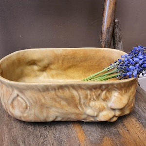May include: A vintage, oblong, tan-colored ceramic planter with embossed floral designs. The planter holds a bunch of blue grape hyacinth flowers. The planter sits on a wooden chair.