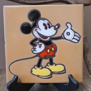 May include: Square ceramic tile featuring a cartoon image of Mickey Mouse. The character is depicted in his classic outfit with black, red, yellow, and white colors. The tile is set on a black display stand.