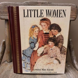 May include: A hardcover book titled Little Women by Louisa May Alcott, illustrated by Jessie Willcox Smith. The cover features a color illustration of four young people in period clothing, with a dark brown spine and gold decorative detailing.