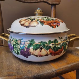 May include: A white enamel cooking pot with a lid, adorned with fruit illustrations. The pot features gold-colored handles and a gold-toned knob on the lid. The fruit designs include pears, grapes, and cherries. The pot is sitting on a wooden chair.