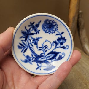 May include: A small, round, white ceramic dish with a blue floral pattern. The design features stylized flowers and foliage. The dish is held in a hand, showcasing its size and detail. The rim is outlined in blue.