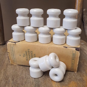 May include: A collection of white ceramic electrical insulators with a spool-like shape, featuring a central groove and a hole through the middle. These cylindrical components are arranged in a group, with some resting on a tan book cover.