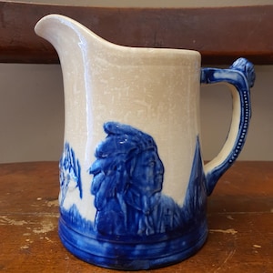 May include: Cream-colored ceramic pitcher with a blue handle and a blue relief design of a Native American chief. The pitcher has a spout and a detailed handle. The design is repeated around the base of the pitcher.