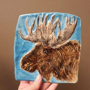 May include: A ceramic tile featuring a detailed relief of a moose head with large antlers. The moose is brown with textured fur, set against a light blue background. The tile is rectangular with rounded corners.