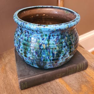 May include: A round ceramic planter featuring a vibrant drip glaze in shades of bright blue, deep navy, and forest green with subtle golden flecks. The exterior has a horizontal ribbed texture, while the interior is a smooth, solid brown finish.