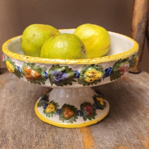 May include: A white ceramic pedestal bowl with a yellow rim and floral designs, holding three green and yellow fruits. The bowl is on a wooden surface, showcasing a rustic aesthetic.