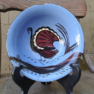 May include: A hand-painted, vintage ceramic bowl with a light blue interior and a black exterior. The bowl features a turkey design with red and black details. The bowl is displayed on a black stand.