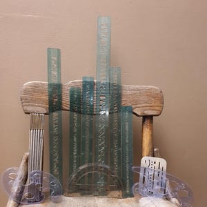 May include: A collection of clear, green-tinted drafting tools, including rulers, stencils, and a protractor, arranged on a wooden chair. The rulers feature etched lettering. The tools are designed for technical drawing and design work.