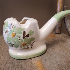 May include: A cream-colored ceramic ashtray shaped like a smoking pipe. The bowl features a painted floral design with light green petals, brown leaves, and yellow centers. The stem has a light green tip, and the base has a green wash.