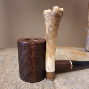May include: A tobacco pipe featuring a cylindrical dark brown wooden bowl with a visible grain pattern. The stem is composed of a textured, light-colored bone-like material connected to a dark brown wooden section and a black mouthpiece with a gold ring.