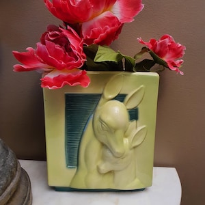 May include: A rectangular, pale yellow ceramic vase with a raised relief of a deer and fawn. The vase has a teal-colored rectangular accent. Bright red flowers with green leaves are arranged in the vase.