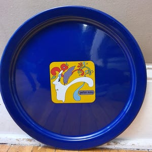May include: A round, cobalt blue metal tray with a glossy finish. The tray features a square, yellow sticker with a white profile, red flower, and the text "peter max".