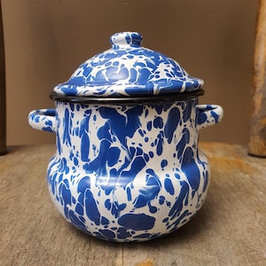 May include: A blue and white enamelware pot with a lid. The pot has a speckled pattern and two small handles. The rim and handles are black. The pot is sitting on a wooden surface.