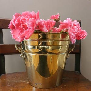 May include: A shiny, gold-colored metal bucket with two handles, filled with vibrant pink flowers. The bucket sits on a wooden chair, creating a classic and elegant display. The flowers add a pop of color to the scene.