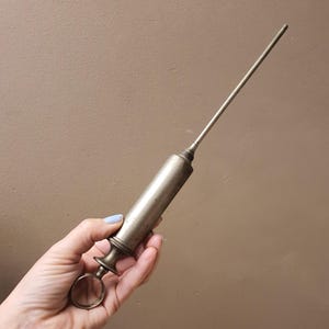 May include: A vintage metallic syringe featuring a cylindrical barrel, a long thin needle, and a circular finger ring at the base of the plunger. The object has a tarnished silver-toned finish and a textured grip area near the plunger handle.
