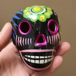 May include: A black ceramic skull figurine featuring hand-painted details in bright colors. The skull has pink eye sockets and a pink nose, a wide mouth with white teeth, and a green and yellow floral pattern on the forehead with gold glitter accents.