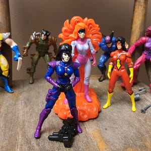 May include: A collection of seven plastic action figures featuring various characters in colorful costumes, including a yellow and blue figure with claws, a green armored figure, a blue and purple figure, and a figure with bright orange hair.
