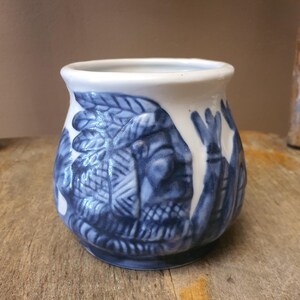May include: A white ceramic mug featuring a blue embossed relief design of a figure wearing a feathered headdress. The mug has a rounded body, a wide opening, and a smooth glazed finish. The blue pattern wraps around the exterior surface.