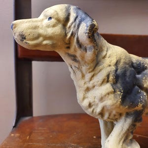May include: A vintage dog figurine in a standing pose, with a cream-colored body and patches of black and gold paint. The dog has a long snout and floppy ears. The figurine is placed on a wooden chair.