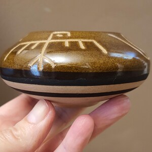 May include: A small, round, ceramic bowl with a brown glaze and a black band. The bowl features a cream-colored geometric design of a bird. The bowl is approximately 3 inches in diameter.