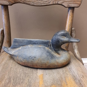 May include: A weathered, cast iron duck planter in shades of blue and gray, resting on a wooden chair. The duck has a detailed head and a trough-like body for planting. The chair has a rustic, aged appearance.