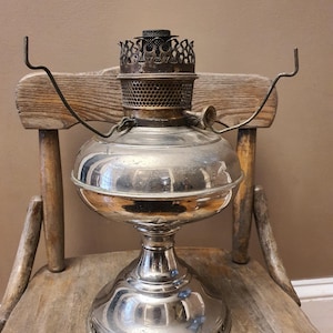 May include: A vintage silver oil lamp with a decorative metal top and a round, bulbous glass reservoir. The lamp has a detailed base and is set on a wooden chair. The lamp's design suggests it is an antique, possibly from the early 20th century.