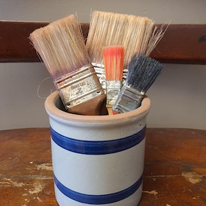May include: A collection of paint brushes of varying sizes and bristle colors, including orange and black, are arranged inside a ceramic crock. The crock is off-white with two blue stripes and sits on a wooden surface.