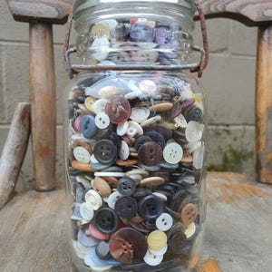 May include: A clear glass jar filled with a variety of buttons in different colors, shapes, and sizes. The buttons are a mix of plastic and other materials. The jar has a metal clasp and a glass lid. The jar is sitting on a wooden chair.