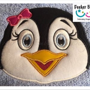 Könnte beinhalten: Gesticktes Pinguin-Gesicht mit einer rosa Schleife, blauen Augen und einem gelben Schnabel. Die Stickerei ist auf einem blauen Stoff.