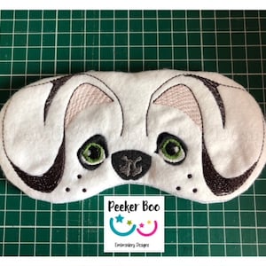 May include: White sleep mask with embroidered black and white dog face. The dog has green eyes and a pink nose. The mask has the text "Peekee Boo Embroidery Designs" on the bottom.