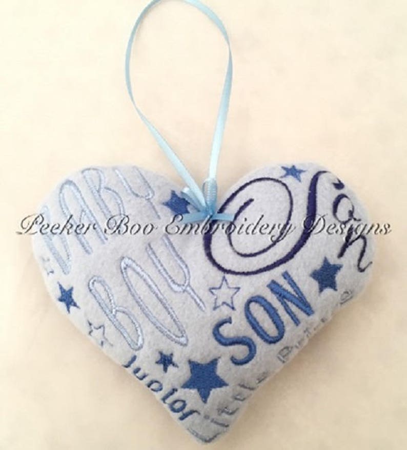 May include: A gray heart-shaped pillow with blue embroidery that reads "Baby Boy" and "Little Son" with stars. The pillow has a blue ribbon attached for hanging.