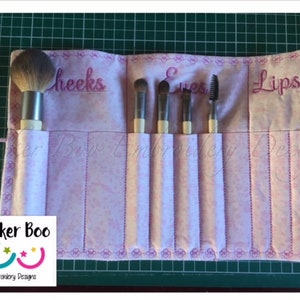 May include: A pink fabric makeup brush roll with embroidered labels for "Cheeks", "Eyes", and "Lips". The roll has six slots for brushes and is rolled up with a pink ribbon.