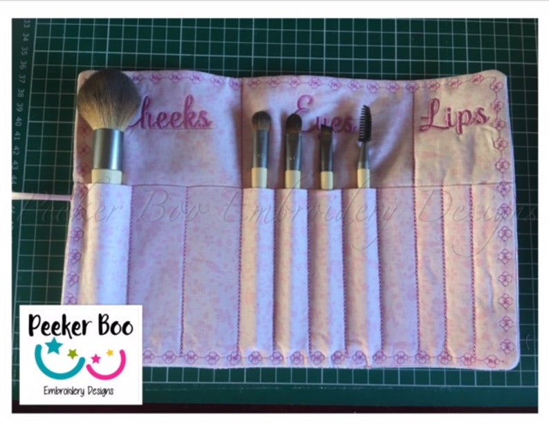 May include: A pink fabric makeup brush roll with embroidered labels for "Cheeks", "Eyes", and "Lips". The roll has six slots for brushes and is rolled up with a pink ribbon.