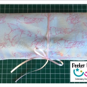 May include: A white fabric with a pink bunny rabbit pattern is rolled up and tied with a pink ribbon. The fabric is labeled "Peeker Boo Embroidery Designs".