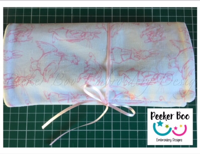 May include: A white fabric with a pink bunny rabbit pattern is rolled up and tied with a pink ribbon. The fabric is labeled "Peeker Boo Embroidery Designs".