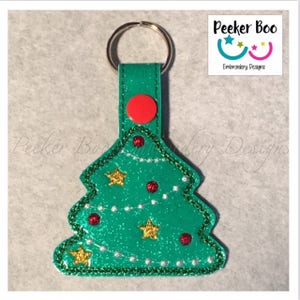 May include: A green glitter Christmas tree keychain with red and white ornaments, gold stars, and a white string of lights. The keychain has a red button and a silver key ring. "Peekee Boo Embroidery Designs" is printed on the top right corner of the image.