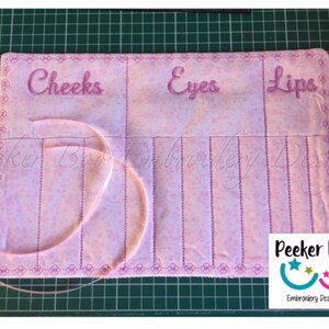 May include: Pink fabric roll-up organizer with sections labeled "Cheeks", "Eyes", and "Lips". The organizer has a pink ribbon and a white label with the text "Peekee Boo Embroidery Designs".
