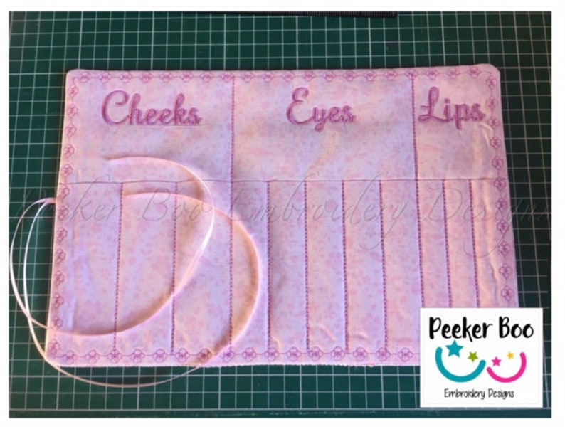 May include: Pink fabric roll-up organizer with sections labeled "Cheeks", "Eyes", and "Lips". The organizer has a pink ribbon and a white label with the text "Peekee Boo Embroidery Designs".