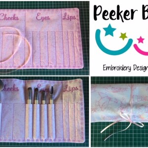 May include: A pink makeup brush roll with embroidered sections labeled "Cheeks", "Eyes", and "Lips". The roll is rolled up and tied with a pink ribbon. The roll is on a green surface. The image also includes a logo for "Peekeer Boo Embroidery Designs" with a blue and pink smiley face design.