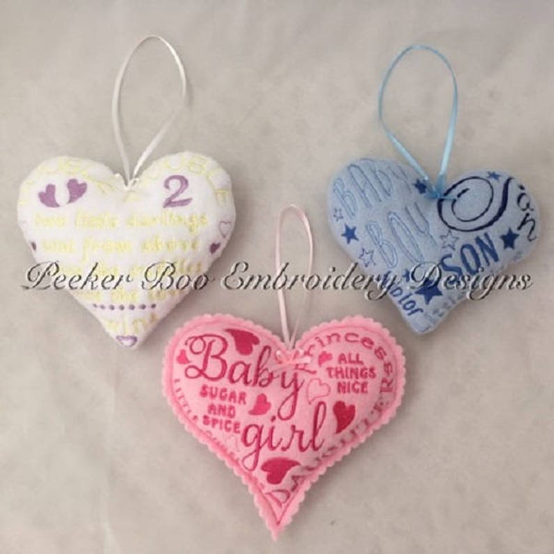 May include: Three felt heart ornaments with embroidered baby-themed designs. The first heart is white with purple and yellow embroidery and the text "New little darling sent from above". The second heart is blue with white and yellow embroidery and the text "Baby Boy Son". The third heart is pink with pink and white embroidery and the text "Baby girl Sugar and Spice".