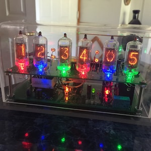 May include: A nixie tube clock featuring six glass vacuum tubes displaying the numbers one through five in glowing orange. The tubes are mounted on a green circuit board with multi-colored LED backlighting, all enclosed in a clear acrylic case.