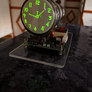 May include: A circular clock with a glowing green face and numerals, mounted on a circuit board base. The clock features a black frame, metal hardware, and exposed electronic components including resistors and capacitors on a clear rectangular platform.