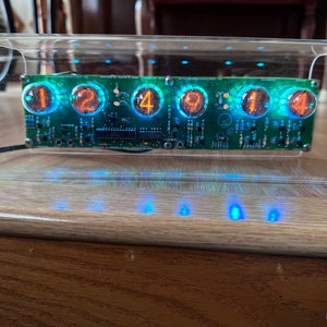 Könnte beinhalten: Ein transparentes Acrylgehäuse mit einer grünen Platine im Inneren. Die Platine hat sechs Nixie-Röhren, die die Zahlen 12:49:14 anzeigen. Das Gehäuse ist mit blauen LEDs beleuchtet.