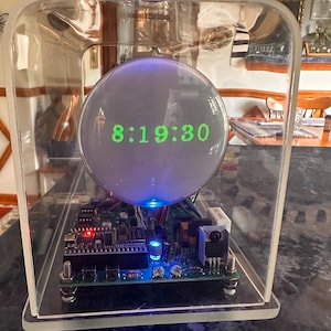 Puede incluir: Un reloj digital con una pantalla de cristal esférica que muestra la hora verde 8:19:30, alojado en una caja rectangular de acrílico transparente. La placa de circuito interna es visible, con componentes electrónicos, luz roja y azul.
