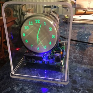 Könnte beinhalten: Eine Uhr mit einem klaren Acrylgehäuse und einem grünen digitalen Display. Die Uhr ist auf 12:30 eingestellt und wird mit blauen LEDs beleuchtet.